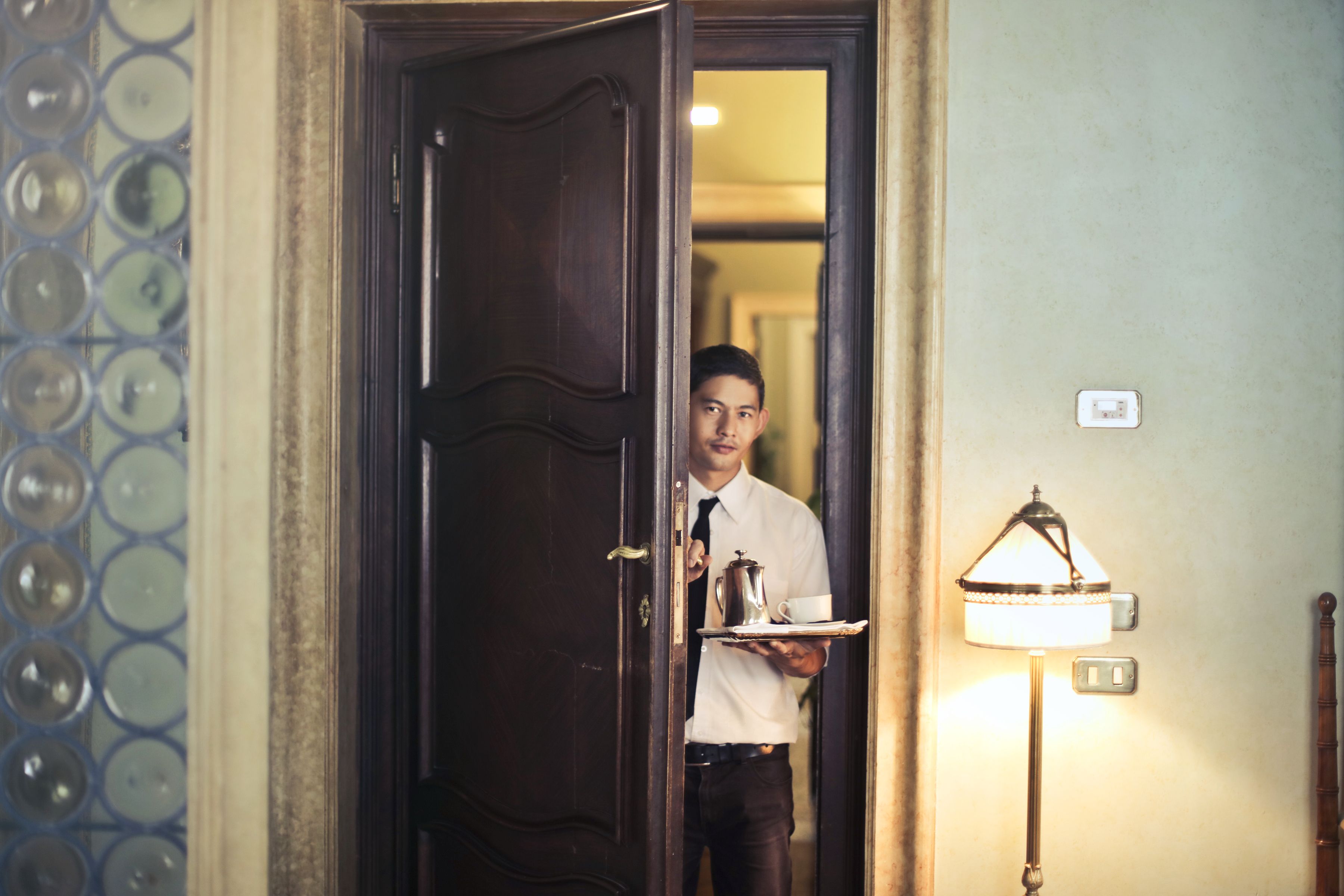 A room-service waiter carries a coffee tray into an elegantly furnished hotel room.